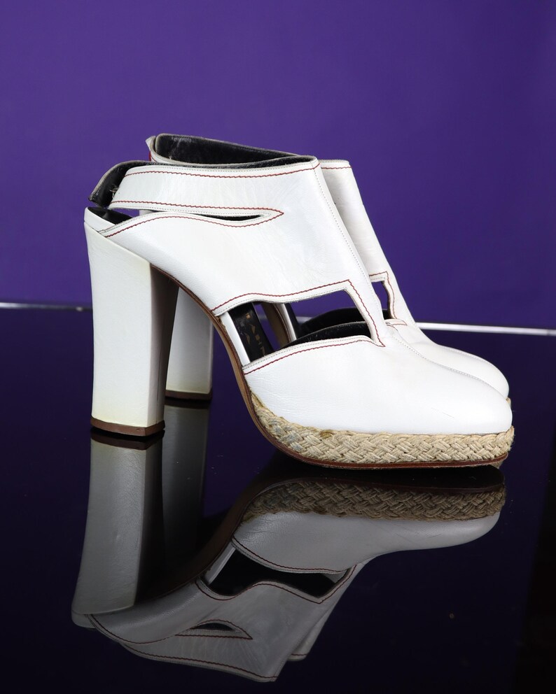May include: White leather high-heeled sandals with cut-out details and a woven jute platform. The sandals have a thick heel and are accented with red stitching. The shoes are reflected on a dark, glossy surface.