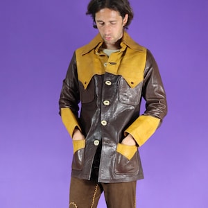 May include: A brown leather jacket featuring a mustard yellow pointed collar, matching yellow cuffs, and yellow pocket flaps. The jacket has a button-down front closure with four visible buttons, two chest pockets, and two lower pockets with yellow accents.