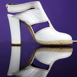 May include: White leather sandal with a thick heel and open design. The sandal features red stitching and a woven jute sole. The shoe has a strap around the ankle and a small red accent. The shoe is reflected on a purple surface.