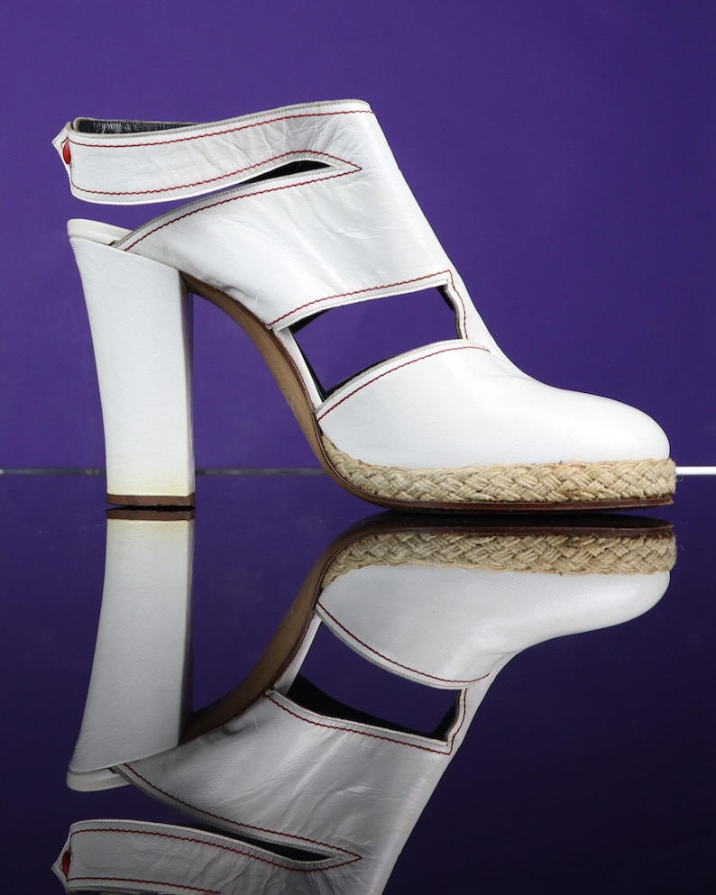 May include: White leather sandal with a thick heel and open design. The sandal features red stitching and a woven jute sole. The shoe has a strap around the ankle and a small red accent. The shoe is reflected on a purple surface.