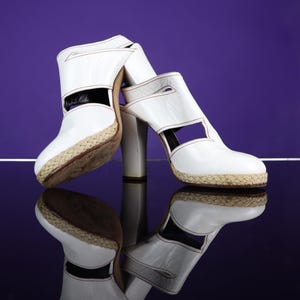 May include: A pair of white leather high-heeled sandals with a woven platform sole. The sandals have a cut-out design and are reflected on a glossy surface against a purple background. The heels are approximately 4 inches tall.