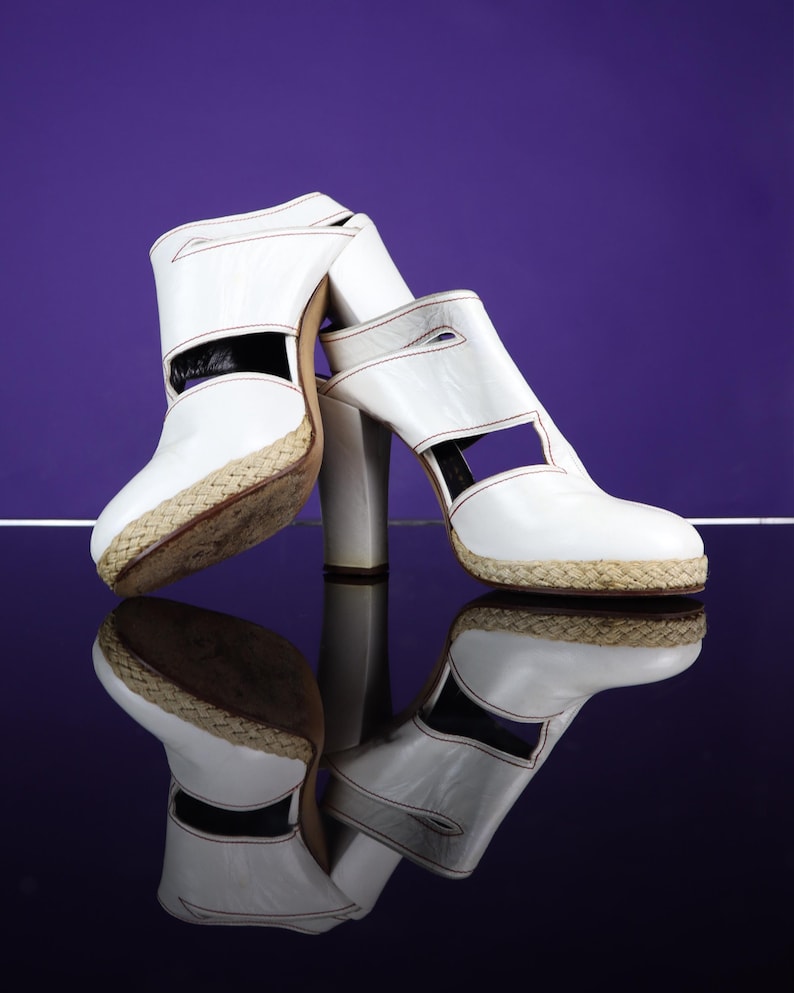 May include: A pair of white leather high-heeled sandals with a woven platform sole. The sandals have a cut-out design and are reflected on a glossy surface against a purple background. The heels are approximately 4 inches tall.