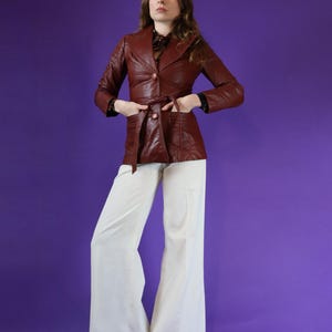 May include: A brown leather jacket with a belt and a single button closure. The jacket has a collar and long sleeves. The model is wearing white wide-leg pants.