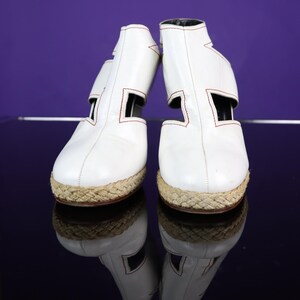May include: A pair of white leather espadrille-style shoes with red stitching and woven jute soles. The shoes have a unique cut-out design on the sides and a black interior. The shoes are reflected on a glossy surface.