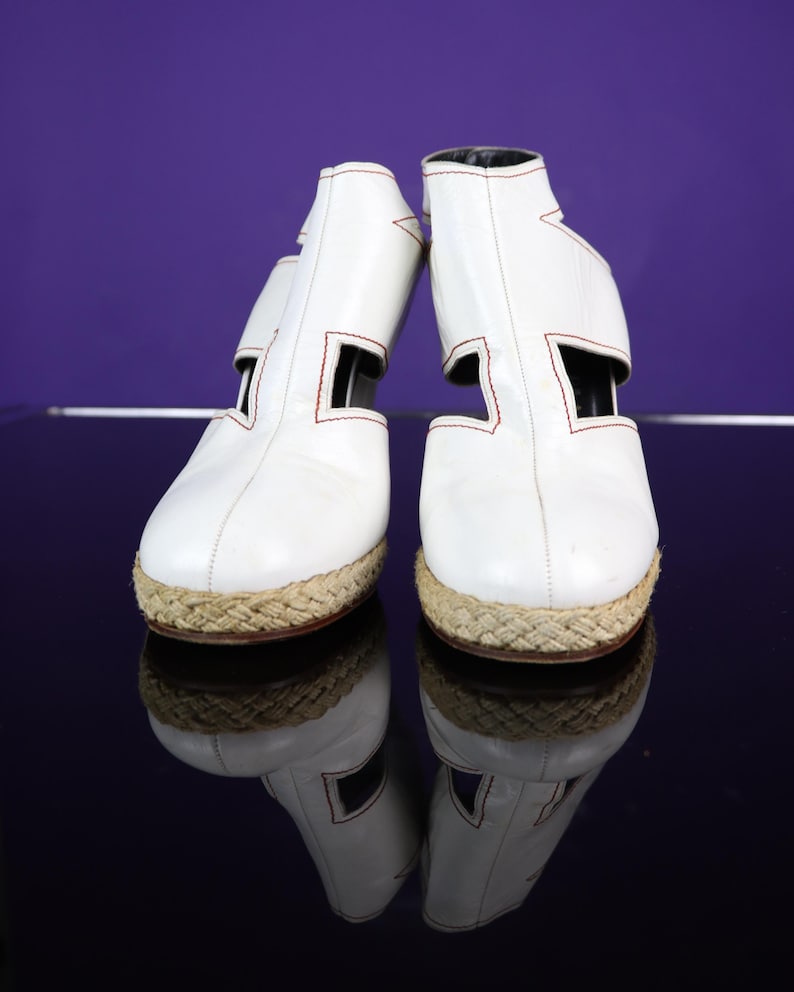 May include: A pair of white leather espadrille-style shoes with red stitching and woven jute soles. The shoes have a unique cut-out design on the sides and a black interior. The shoes are reflected on a glossy surface.