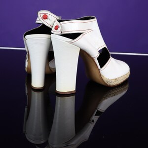 May include: White leather high-heeled sandals with red stitching and accents. The sandals feature a chunky heel, open sides, and a woven platform sole. The design includes a strap with a red button closure. The shoes are reflected on a dark surface.