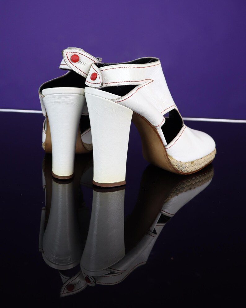 May include: White leather high-heeled sandals with red stitching and accents. The sandals feature a chunky heel, open sides, and a woven platform sole. The design includes a strap with a red button closure. The shoes are reflected on a dark surface.