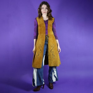 May include: A long brown suede waistcoat with a tie closure and snake embroidery. The waistcoat is worn over a purple shirt and blue jeans.