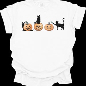 May include: White t-shirt featuring a Halloween design. The graphic shows three carved pumpkins with smiling faces and black cats. One cat sits atop a pumpkin, while others interact with the pumpkins. The design is in black and orange.