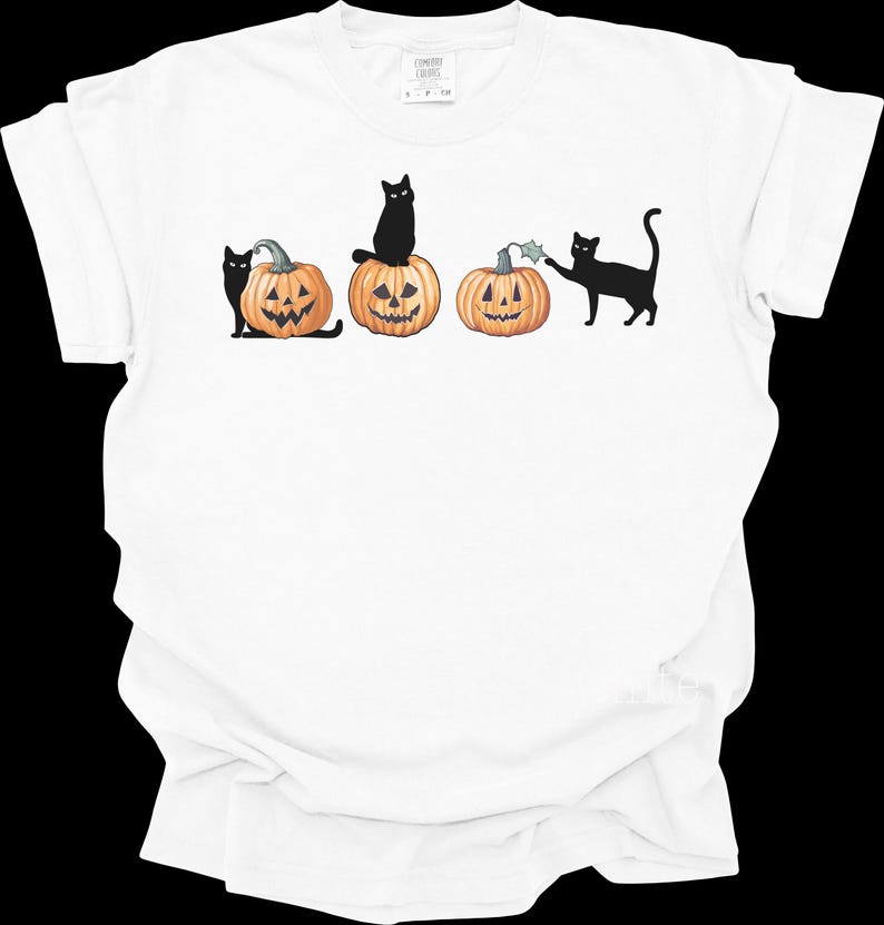 May include: White t-shirt featuring a Halloween design. The graphic shows three carved pumpkins with smiling faces and black cats. One cat sits atop a pumpkin, while others interact with the pumpkins. The design is in black and orange.