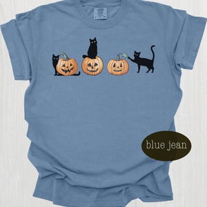 May include: Blue jean colored t-shirt featuring a Halloween design. The graphic includes black cats interacting with carved jack-o'-lanterns. The shirt has a crew neck and short sleeves. The text "blue jean" is on the bottom right.