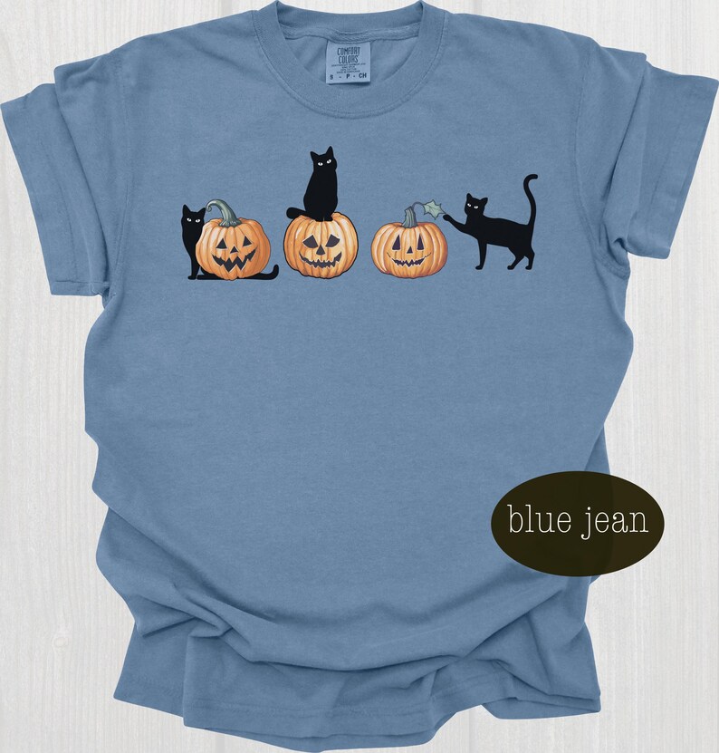 May include: Blue jean colored t-shirt featuring a Halloween design. The graphic includes black cats interacting with carved jack-o'-lanterns. The shirt has a crew neck and short sleeves. The text "blue jean" is on the bottom right.