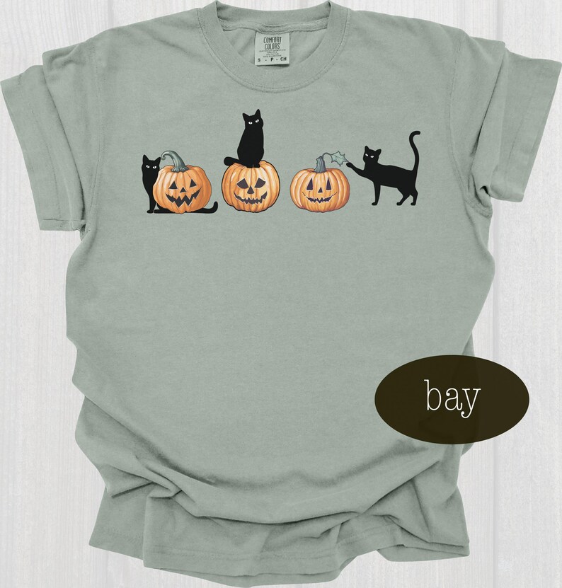 May include: A light green t-shirt featuring a Halloween design. The design includes three carved pumpkins with black cats. One cat sits atop a pumpkin, another stands beside a pumpkin, and the third interacts with a pumpkin.