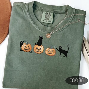 May include: Moss green t-shirt featuring a graphic of three jack-o'-lanterns with black cats. The shirt has a Comfort Colors tag and a small gold necklace. The word "moss" is printed on the bottom right.