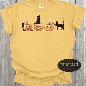 May include: Mustard-colored t-shirt featuring a Halloween design. The graphic includes three carved pumpkins and black cats. One cat sits atop a pumpkin, another interacts with a pumpkin, and the third sits beside a pumpkin. The shirt has a crew neck.