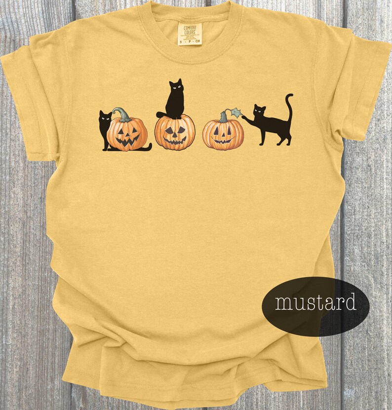 May include: Mustard-colored t-shirt featuring a Halloween design. The graphic includes three carved pumpkins and black cats. One cat sits atop a pumpkin, another interacts with a pumpkin, and the third sits beside a pumpkin. The shirt has a crew neck.