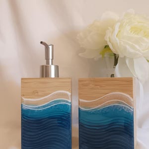 Op de afbeelding: Een bamboe zeepdispenser met een roestvrijstalen pomp en een bijpassende rechthoekige houder. De voorkant van elk item heeft een gelaagd ontwerp van wit, beige en blauwtinten, dat op oceaangolven lijkt.