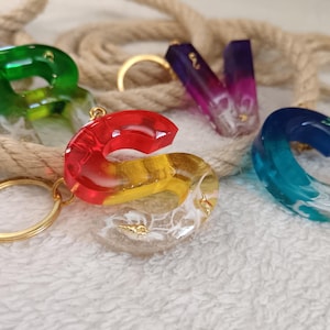 Può includere: Una collezione di portachiavi in resina colorata in varie forme, tra cui le lettere "S" e "V". I portachiavi presentano elementi dorati e sono riempiti di glitter ed elementi decorativi. Una corda è sullo sfondo.