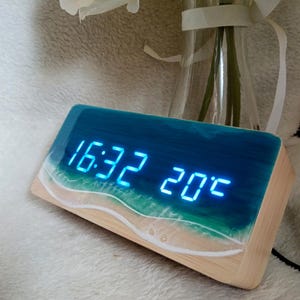 Può includere: Un orologio digitale in legno con display a LED blu che mostra l'ora e la temperatura. La parte superiore dell'orologio presenta un design in resina che raffigura una scena di spiaggia con tonalità blu e verdi. L'orologio mostra 16:32 e 20°C.