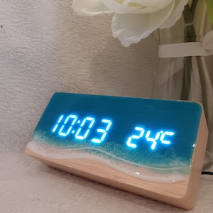 Op de afbeelding: Een rechthoekige digitale wekker met een lichte houten basis en een hars bovenkant met een blauw oceaangolfontwerp met wit schuim. De wijzerplaat toont de felblauwe digitale cijfers 10:03 en de temperatuur 24C op het vooroppervlak.