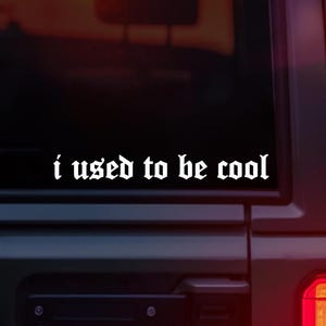 Może przedstawiać: Biała naklejka winylowa z napisem "i used to be cool" w gotyckiej czcionce. Naklejka jest umieszczona na tylnej szybie pojazdu, a światło tylne jest widoczne w prawym dolnym rogu.