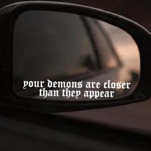 May include: A black car wing mirror with the text "your demons are closer than they appear" in a gothic font. The mirror reflects a blurred outdoor scene.