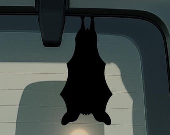 Könnte beinhalten: Ein schwarzer, fledermausförmiger Aufkleber, der kopfüber hängt, ist an der Heckscheibe eines Fahrzeugs angebracht. Die Silhouette des Aufklebers ist deutlich gegen die Scheibe zu sehen. Das Bild hat ein Halloween- oder Gruselthema.