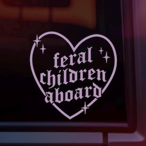 May include: A light purple heart-shaped sticker with the words "feral children aboard" in a gothic font. The sticker has four star accents and is affixed to a vehicle window.