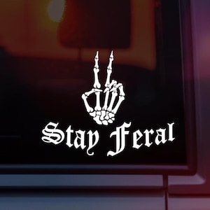 May include: White decal showing a skeleton hand in a peace sign gesture. The words "Stay Feral" are written in a gothic font beneath the hand. The decal is on a dark background.