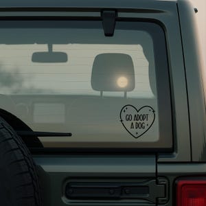 May include: Black heart-shaped car decal with the text "GO ADOPT A DOG +" in a sans-serif font. The decal is affixed to the rear window of a dark green vehicle. The image is taken outdoors.