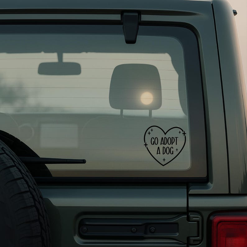May include: Black heart-shaped car decal with the text "GO ADOPT A DOG +" in a sans-serif font. The decal is affixed to the rear window of a dark green vehicle. The image is taken outdoors.