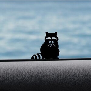 May include: A black raccoon silhouette with white accents, featuring a striped tail and a seated pose. The raccoon is set against a blurred blue and white background, creating a simple yet striking design.