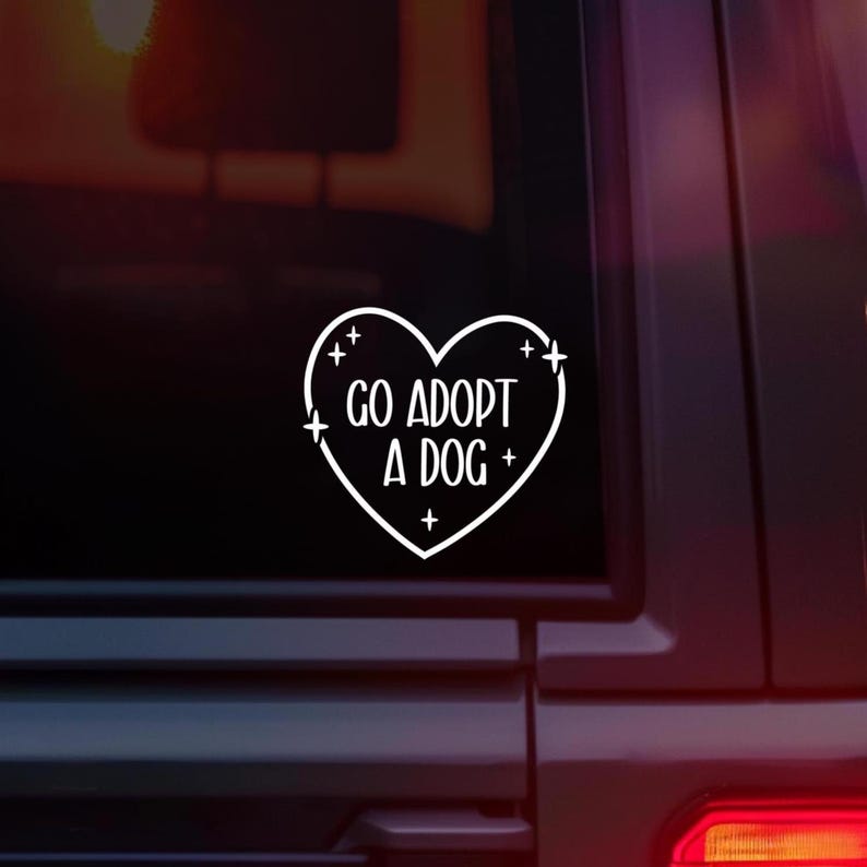 May include: White heart-shaped sticker with the text "GO ADOPT A DOG" in a handwritten font. The sticker has small star accents and is displayed on a dark background, likely a car window.