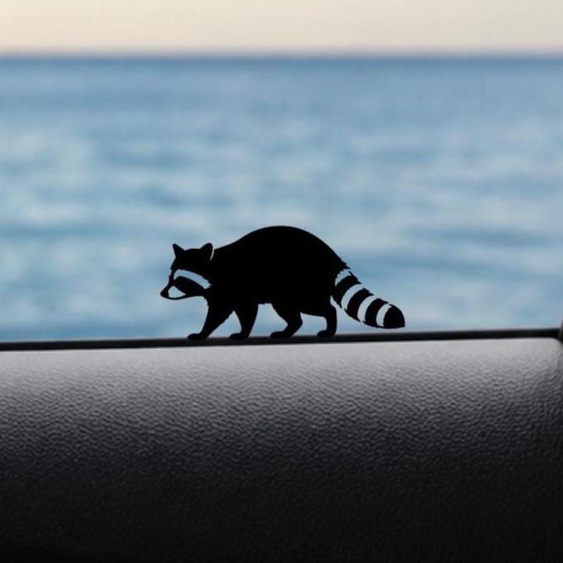 May include: A black raccoon silhouette with white markings walks along a dark surface. The raccoon has a striped tail and a bandit mask. The background features a blurred view of the ocean and sky.