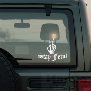 May include: White vinyl decal featuring a skeleton hand making a peace sign, with the words "Stay Feral" in gothic font. The decal is applied to the rear window of a vehicle, with a view of the interior and a bright light visible through the glass.