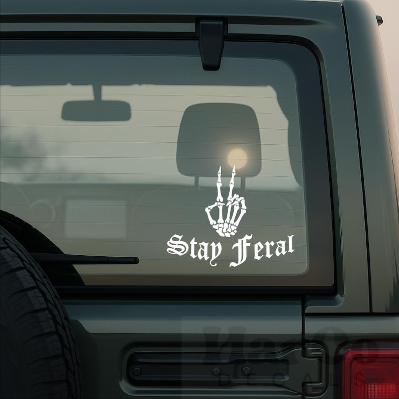 May include: White vinyl decal featuring a skeleton hand making a peace sign, with the words "Stay Feral" in gothic font. The decal is applied to the rear window of a vehicle, with a view of the interior and a bright light visible through the glass.
