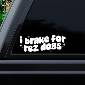 May include: White vinyl decal with the text "i brake for rez dogs" in a playful, rounded font. The decal is affixed to a car window, with a dark background. Small cross symbols are placed around the text.