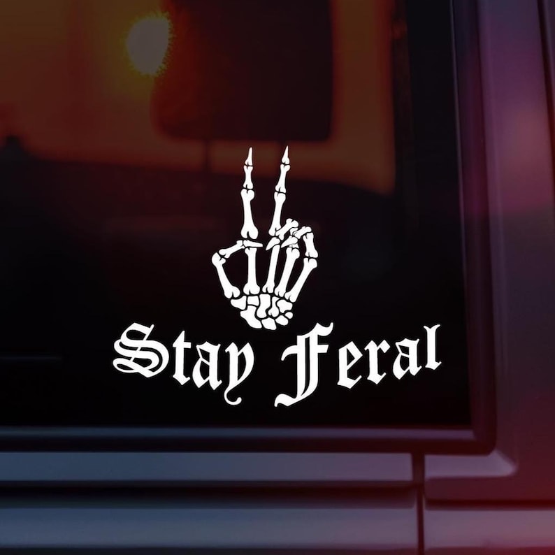 May include: White decal showing a skeleton hand in a peace sign gesture. The words "Stay Feral" are written in a gothic font beneath the hand. The decal is on a dark background.