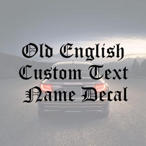 Peut inclure: Une voiture noire avec le texte "Old English Custom Text Name Decal" en police gothique. La voiture est vue de l'arrière, avec ses feux allumés. L'arrière-plan est un paysage flou.