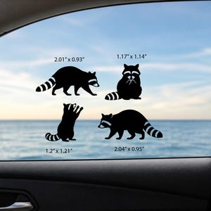 May include: Four black raccoon decals in various poses are displayed on a car window. The decals measure 2.01" x 0.93", 1.17" x 1.14", 1.2" x 1.21", and 2.04" x 0.95". The background shows a blue sky and ocean.
