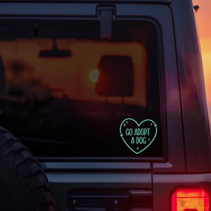 May include: A teal heart-shaped sticker on a car window with the words "GO ADOPT A DOG" in a sans-serif font. The sticker is outlined with a dotted line and small crosses. The background is a sunset.