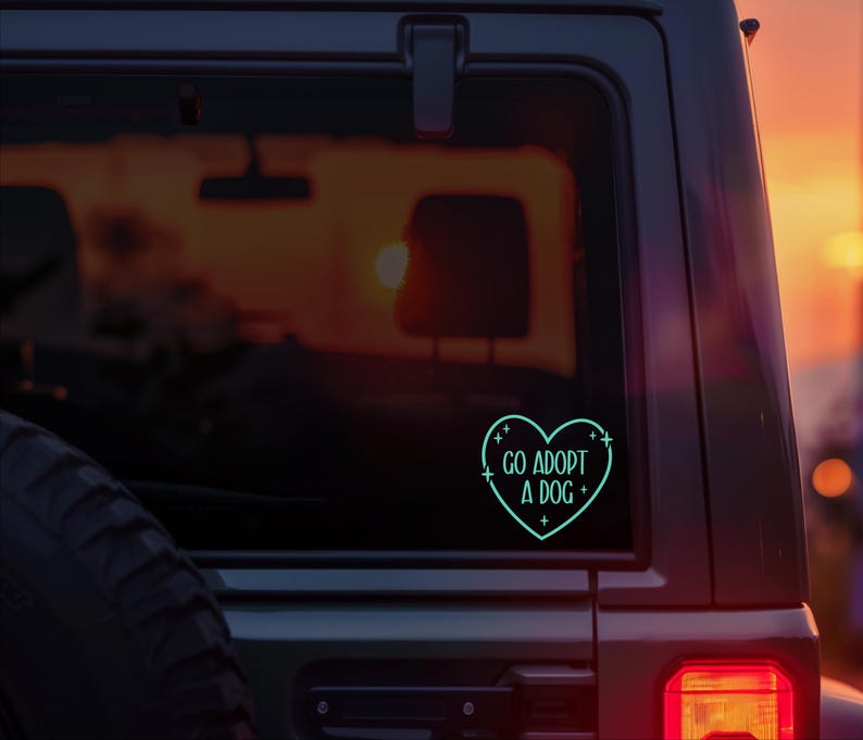May include: A teal heart-shaped sticker on a car window with the words "GO ADOPT A DOG" in a sans-serif font. The sticker is outlined with a dotted line and small crosses. The background is a sunset.