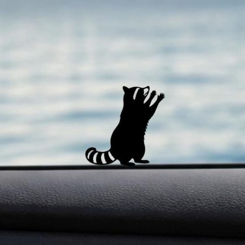 May include: Black raccoon silhouette with a striped tail and raised paws. The raccoon is standing upright, with its arms raised. The background is a blurred, light blue and white. The raccoon is a sticker or decal.