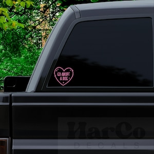 May include: A pink heart-shaped decal with the words "GO ADOPT A DOG" in a handwritten font, with small star accents. The decal is affixed to the window of a black vehicle, with a green leafy background.