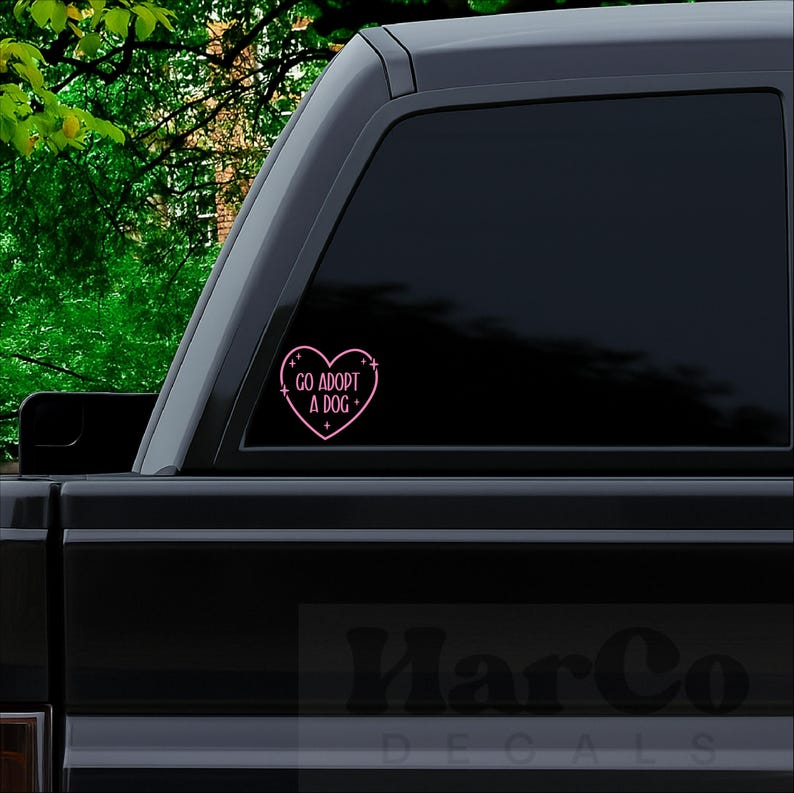 May include: A pink heart-shaped decal with the words "GO ADOPT A DOG" in a handwritten font, with small star accents. The decal is affixed to the window of a black vehicle, with a green leafy background.