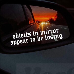 Può includere: Specchietto retrovisore per auto con la scritta "objects in mirror appear to be losing" in bianco, con carattere gotico. Lo specchietto riflette un tramonto con sfumature arancioni e gialle e la strada.