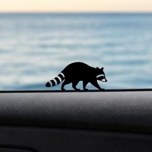 May include: A black raccoon silhouette decal with a striped tail and white accents. The raccoon is walking on a dark surface, with a blurred blue and white background. This decal is suitable for car windows or other smooth surfaces.