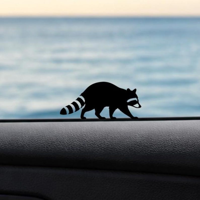 May include: A black raccoon silhouette decal with a striped tail and white accents. The raccoon is walking on a dark surface, with a blurred blue and white background. This decal is suitable for car windows or other smooth surfaces.