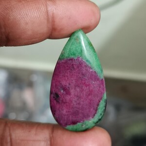 Peut inclure: Une pierre précieuse en zoïsite en forme de goutte d'eau présentant une inclusion de rubis magenta texturée et proéminente en son centre, entourée d'une matrice de zoïsite vert vif avec de subtiles taches minérales sombres sur la surface polie.