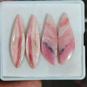 May include: Four polished pink opal gemstones arranged in a white display box. Two stones are elongated marquise shapes with vertical reddish-pink streaks, while the other two are teardrop shapes featuring soft pink hues and dark inclusions.